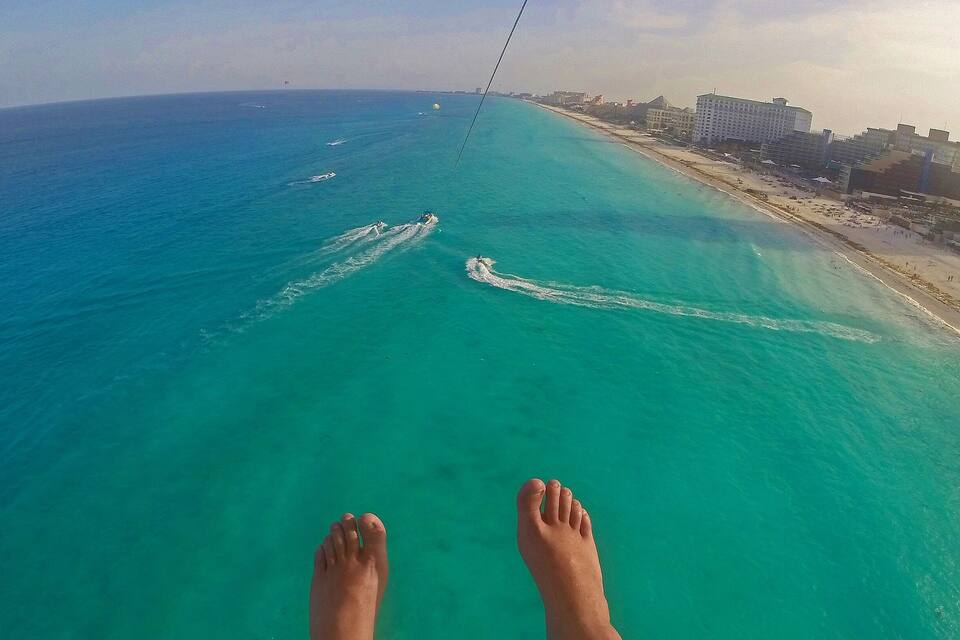 A pair of feet dangling over turquoise ocean water with a beach and buildings in the background.