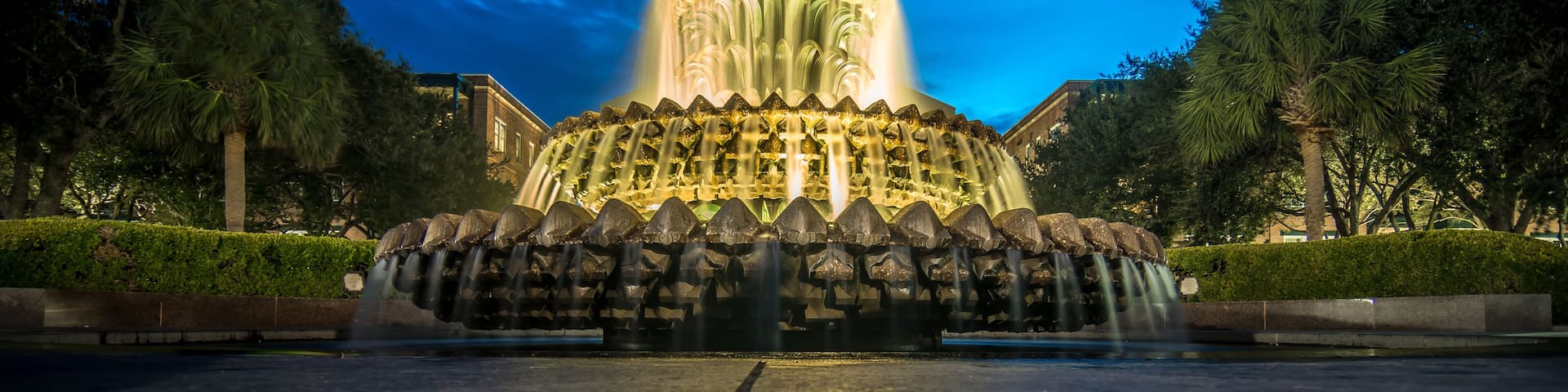 A nighttime illuminated fountain with a pineapple-like top, surrounded by trees and a building in the background.