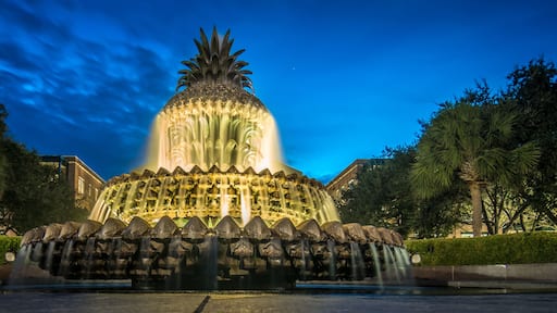 Una fuente iluminada de noche con una parte superior similar a una piña, rodeada de árboles y un edificio al fondo.