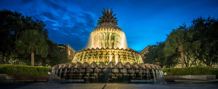 Uma fonte iluminada à noite, com um topo semelhante a uma abacaxi, rodeada por árvores e um edifício ao fundo.