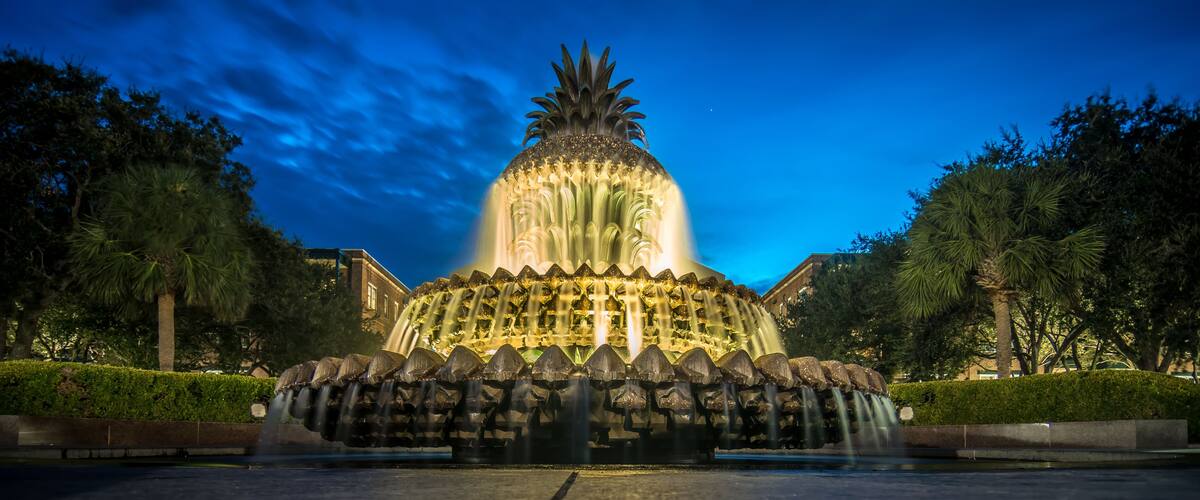 A nighttime illuminated fountain with a pineapple-like top, surrounded by trees and a building in the background.