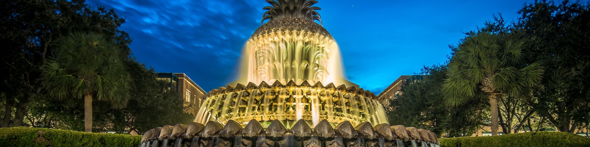 A nighttime illuminated fountain with a pineapple-like top, surrounded by trees and a building in the background.