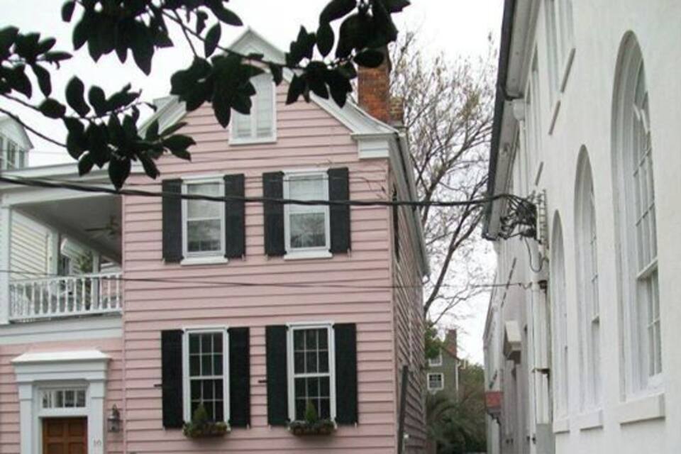 A pink house with black shutters, a white building with arched windows, and a sidewalk.
