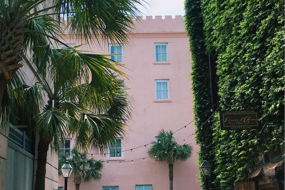 A narrow alley with a pink building, palm trees, and a parked car.