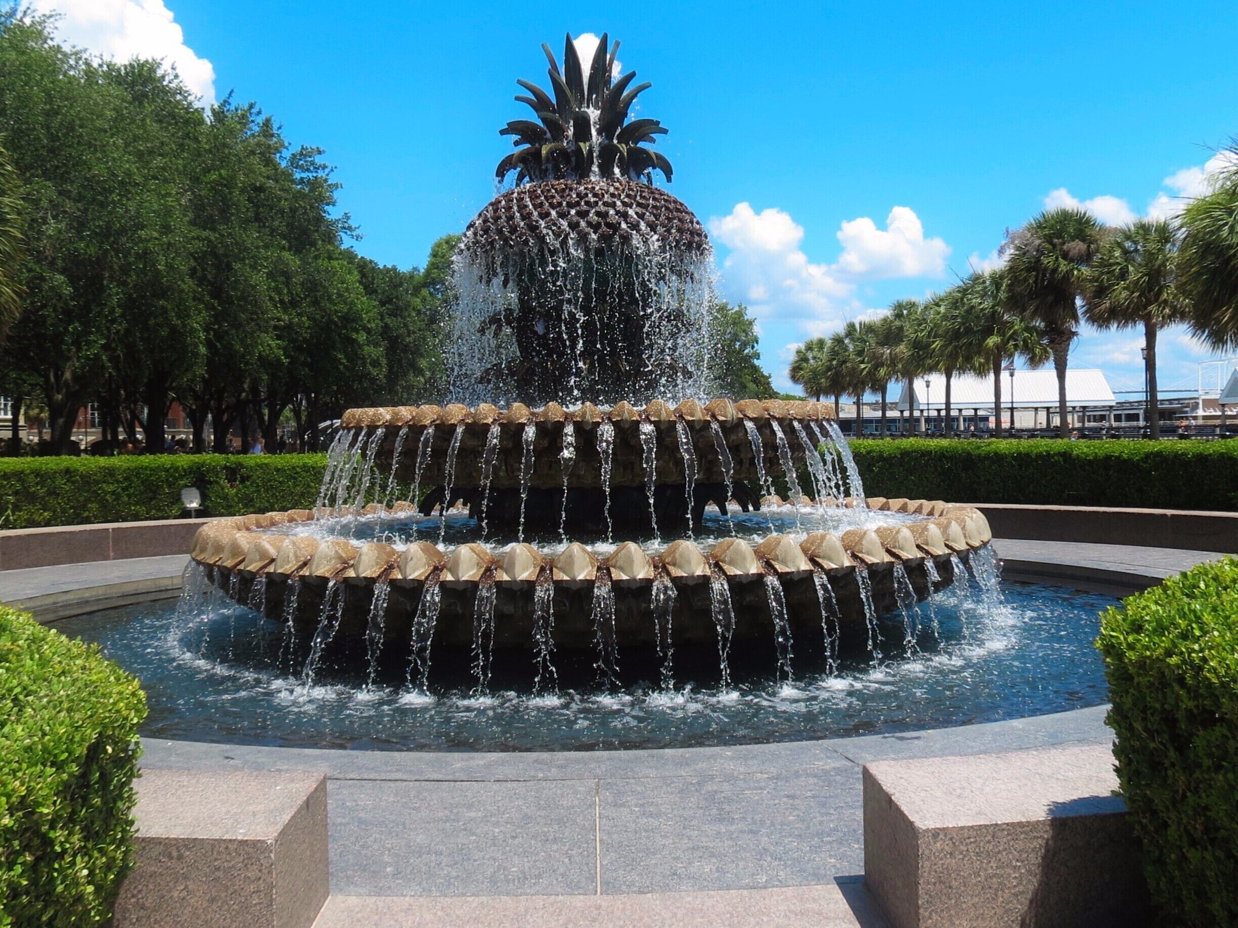 A tiered water fountain with a central statue, surrounded by well-manicured bushes and palm trees.