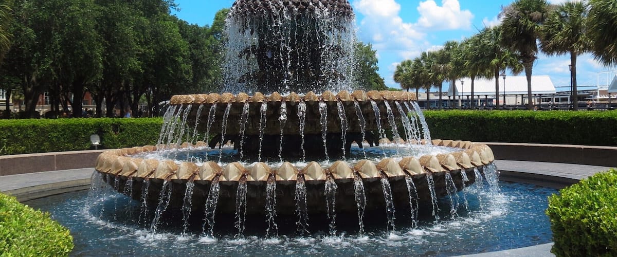 A tiered water fountain with a central statue, surrounded by well-manicured bushes and palm trees.