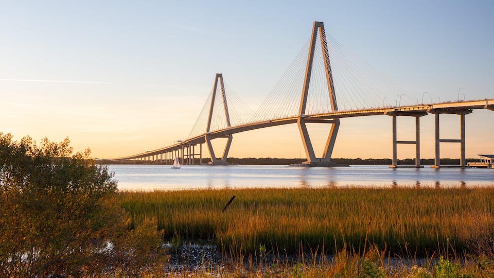A large suspension bridge with tall towers and cables, spanning a body of water with a grassy shoreline in the foreground.