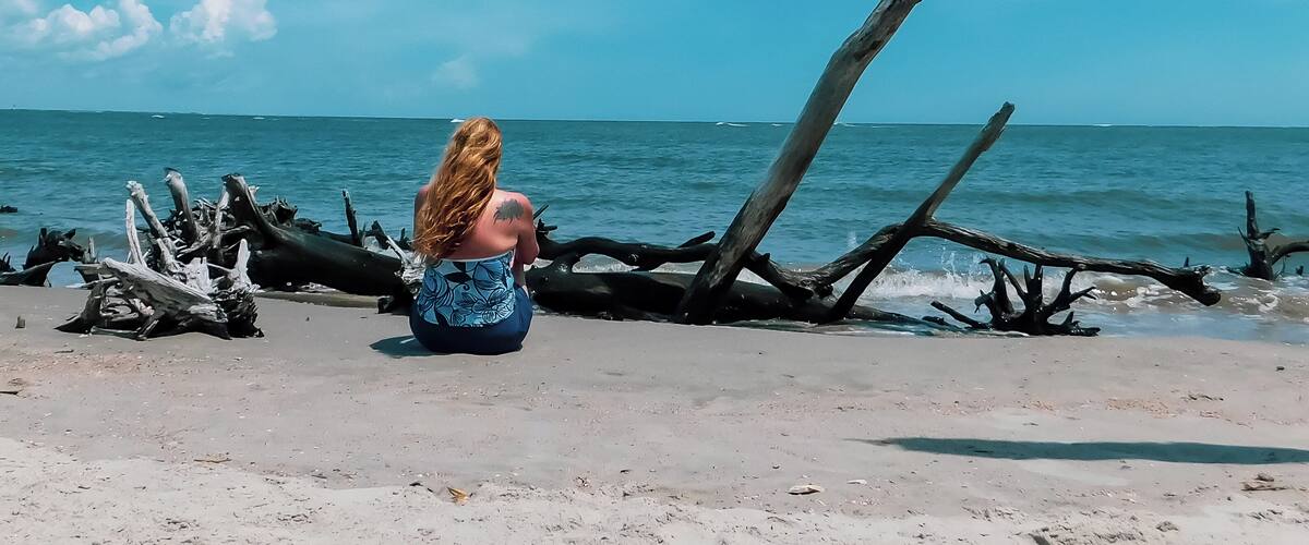 A woman sitting on a beach with driftwood and the ocean in the background.
