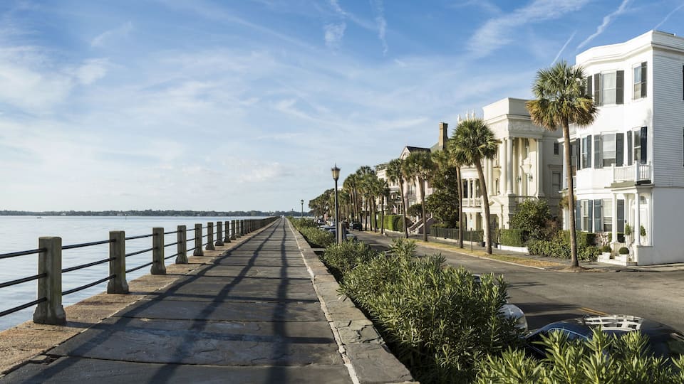 A waterfront promenade with a wooden railing, a paved walkway, and white buildings with shutters.
