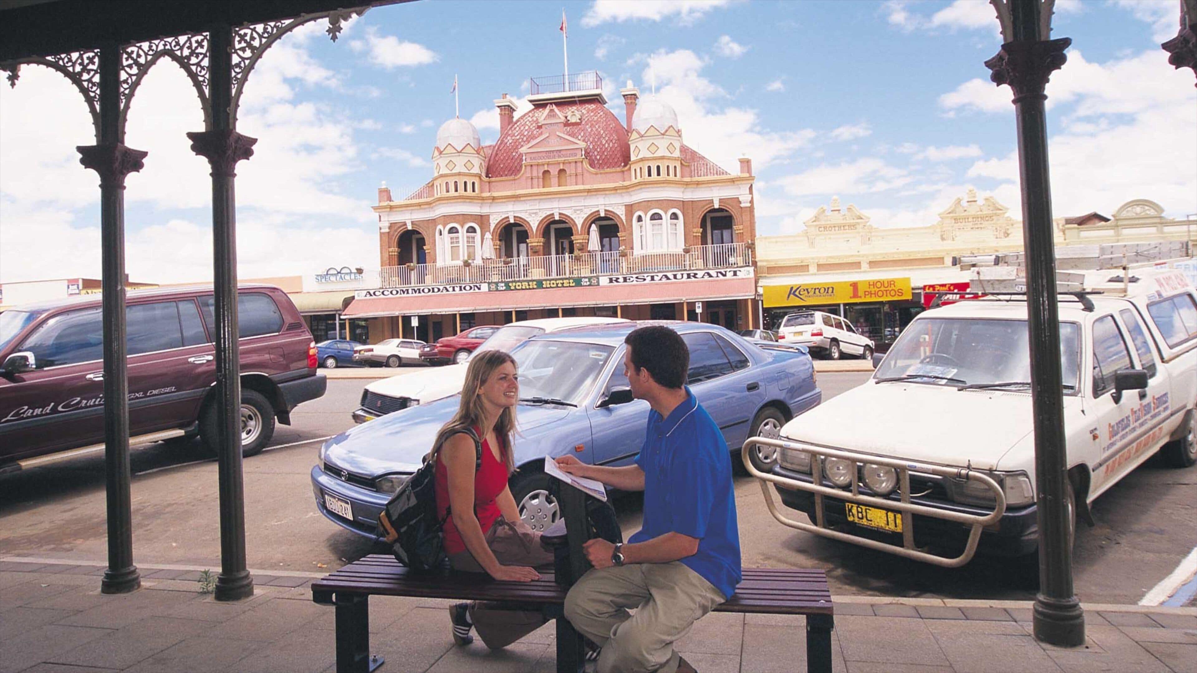 A historic building with a dome and arched windows, surrounded by parked cars and a pedestrian walkway.