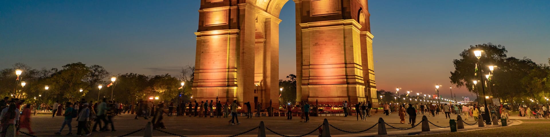 The illuminated India Gate at sunset with people around it.