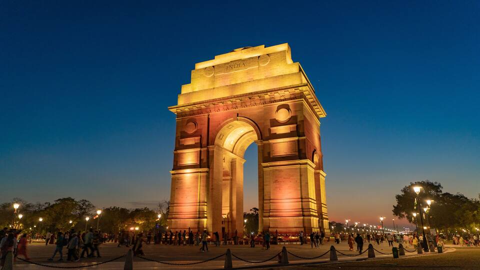 The illuminated India Gate at sunset with people around it.