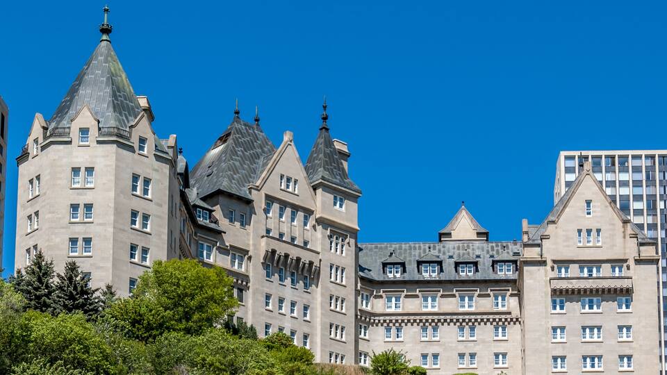 A large, historic building with multiple towers and a steep roof, surrounded by greenery.