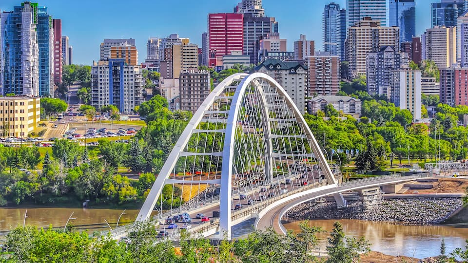 A modern bridge with a white arch structure, surrounded by a cityscape with various buildings and a river.