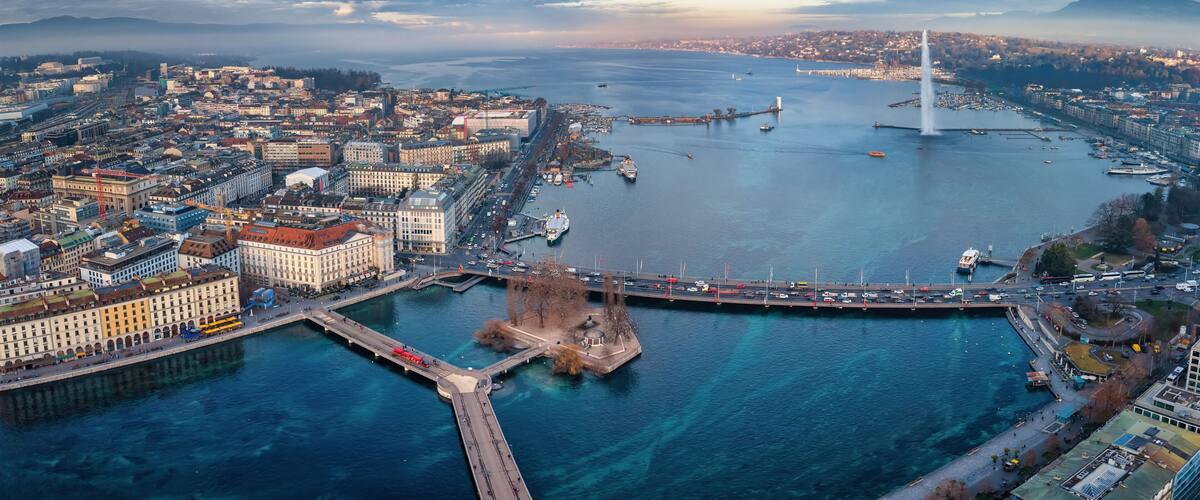Un paysage urbain côtier avec un pont imposant, une vaste étendue d’eau et une chaîne de montagnes lointaine.