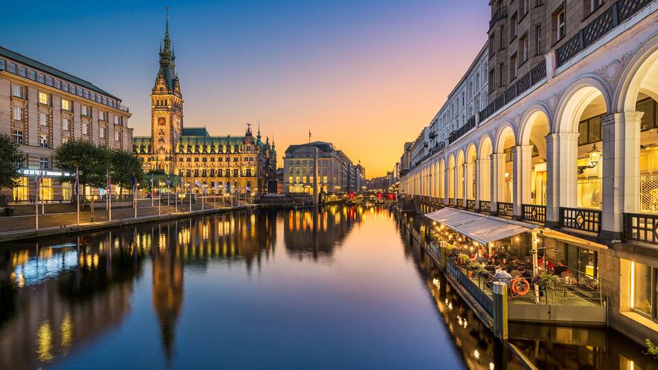 A calm river with buildings on either side, including a prominent tower and a covered walkway.