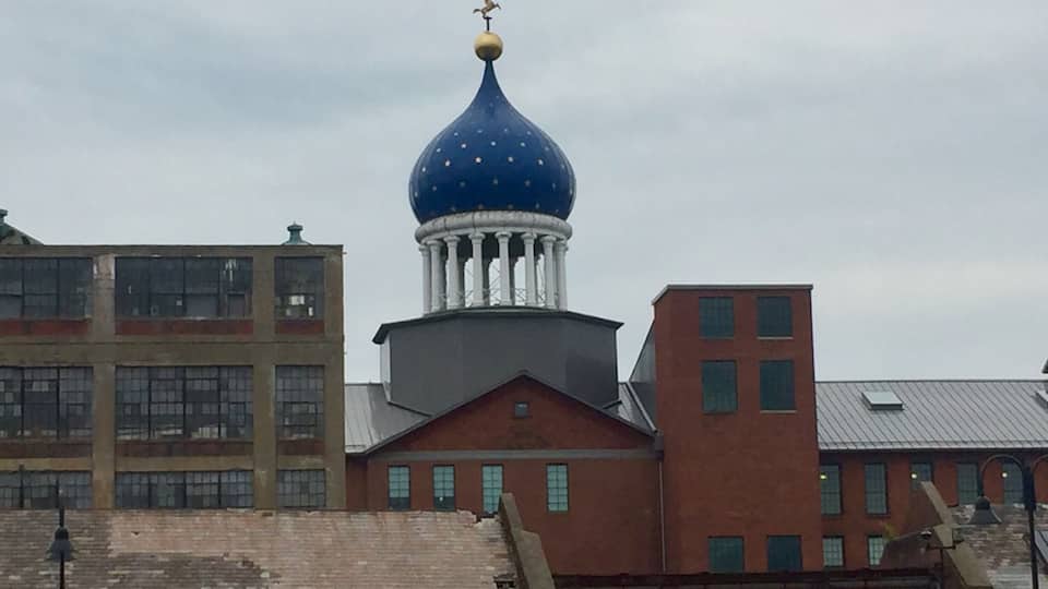 Underneath that blue dome lies the east armory of a factory once owned by a man who changed American history, the innovator of the revolver, Samuel Colt. Sprawling around the crumbling red brickwork of the old Colt’s Patent Firearms Manufacturing Company, once the largest private armory in the world, are the remnants of Coltsville, the utopian village he built for his workers.
A model of 19th century industrial paternalism, Coltsville included a church, a social hall for dances and lectures, workers’ housing, a giant landscaped park home to deer and peacocks, sculpted botanical gardens, and thousands of feet of greenhouses filled with tropical fruit and flowers. Samuel Colt felt such responsibility for the welfare of his workers outside the factory that he went so far as to build Potsdam Village, to replicate the feel of the German village from which he imported his skilled craftsmen to work in his willow ware factory.
The National Park Service began having on-site presence in 2015, with workshops and tours, including up into the Blue Onion Dome which affords magnificent views of downtown Hartford, the Connecticut River and Colt Park, formerly the back yard of Armsmear, Samuel & Elizabeth Colt’s mansion. By 2016, an NPS ranger had taken up residence in the South Armory and was giving walking tours of Coltsville.
The old South, East and North armories are under renovation and the two 1855 brownstone buildings, the oldest structures on site, will become the National Park Service Visitor Center. The armories themselves have been or are being renovated into apartments, offices and education facilities.