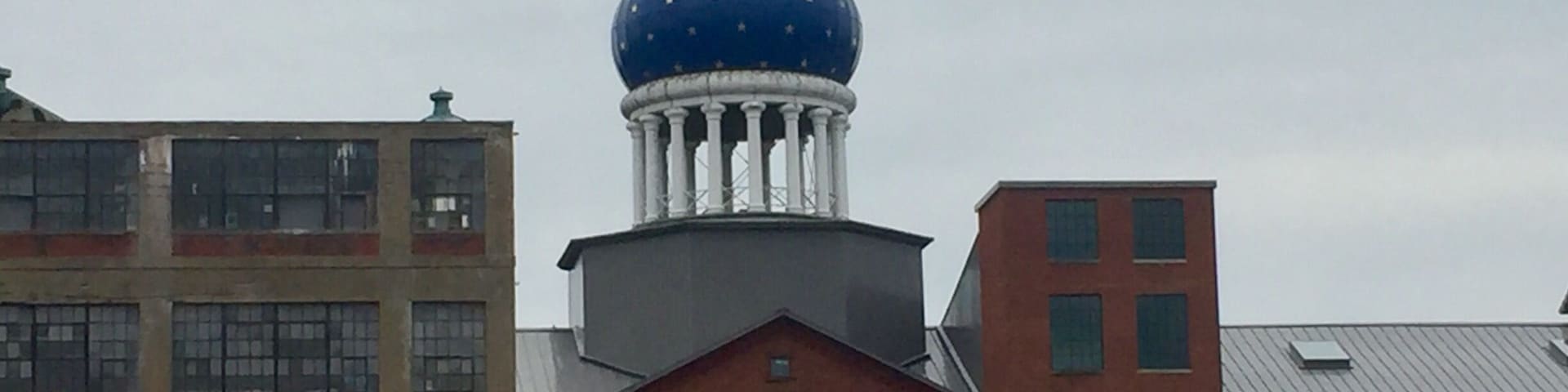 Underneath that blue dome lies the east armory of a factory once owned by a man who changed American history, the innovator of the revolver, Samuel Colt. Sprawling around the crumbling red brickwork of the old Colt’s Patent Firearms Manufacturing Company, once the largest private armory in the world, are the remnants of Coltsville, the utopian village he built for his workers. 
A model of 19th century industrial paternalism, Coltsville included a church, a social hall for dances and lectures, workers’ housing, a giant landscaped park home to deer and peacocks, sculpted botanical gardens, and thousands of feet of greenhouses filled with tropical fruit and flowers. Samuel Colt felt such responsibility for the welfare of his workers outside the factory that he went so far as to build Potsdam Village, to replicate the feel of the German village from which he imported his skilled craftsmen to work in his willow ware factory.
The National Park Service began having on-site presence in 2015, with workshops and tours, including up into the Blue Onion Dome which affords magnificent views of downtown Hartford, the Connecticut River and Colt Park, formerly the back yard of Armsmear, Samuel & Elizabeth Colt’s mansion. By 2016, an NPS ranger had taken up residence in the South Armory and was giving walking tours of Coltsville.
The old South, East and North armories are under renovation and the two 1855 brownstone buildings, the oldest structures on site, will become the National Park Service Visitor Center. The armories themselves have been or are being renovated into apartments, offices and education facilities.