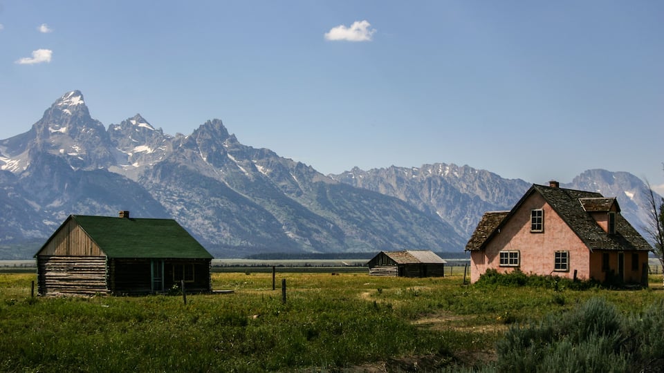 A rural landscape with two wooden houses and a mountain range in the background.