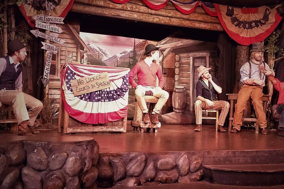 A stage with a rustic backdrop, wooden benches, and a sign reading "No Licker Courts in Shesun."