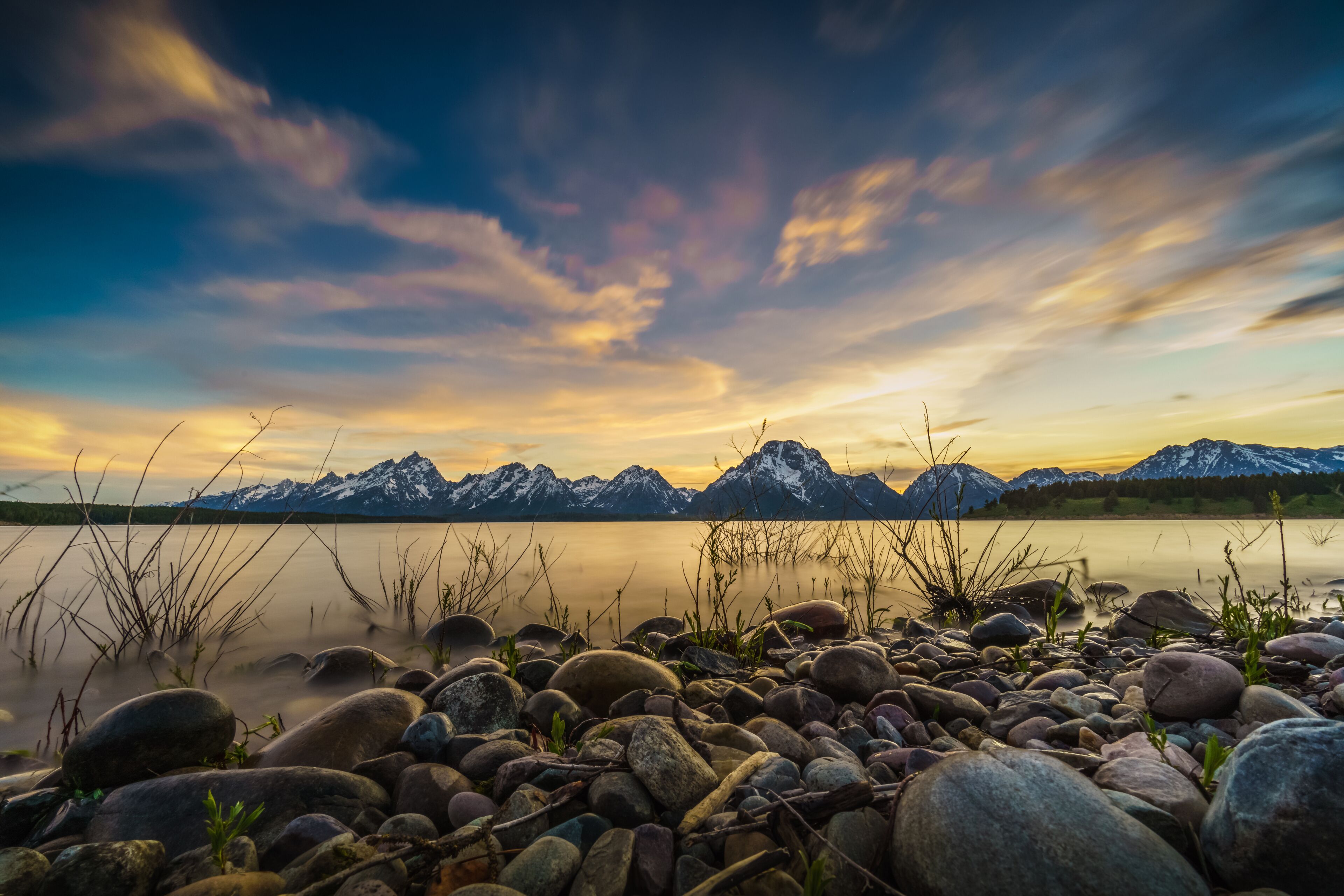 A lake with a rocky shoreline, surrounded by mountains with snow caps, under a colorful sky.