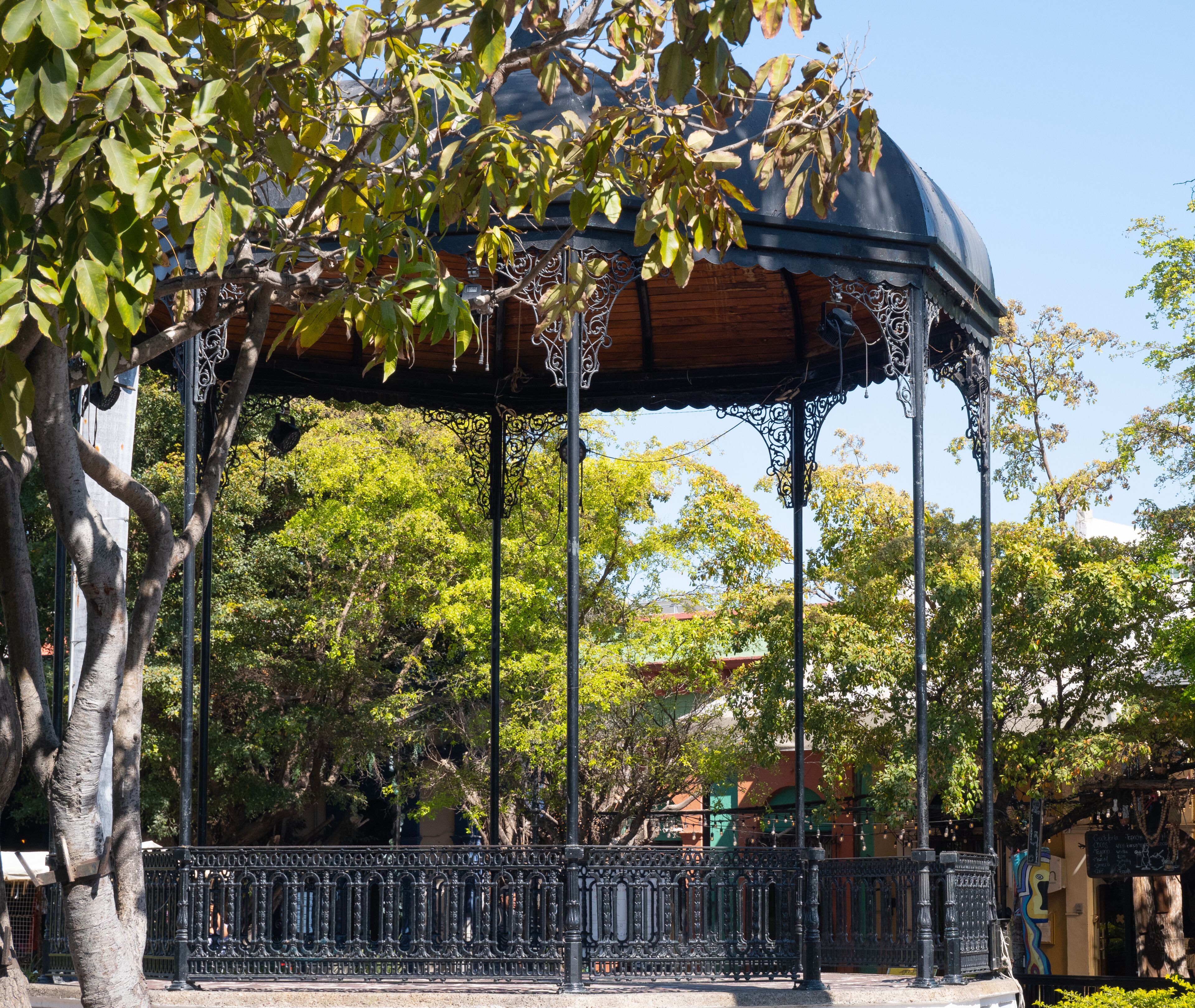 Un pabellón con detalles metálicos ornamentados, rodeado de árboles y una valla.