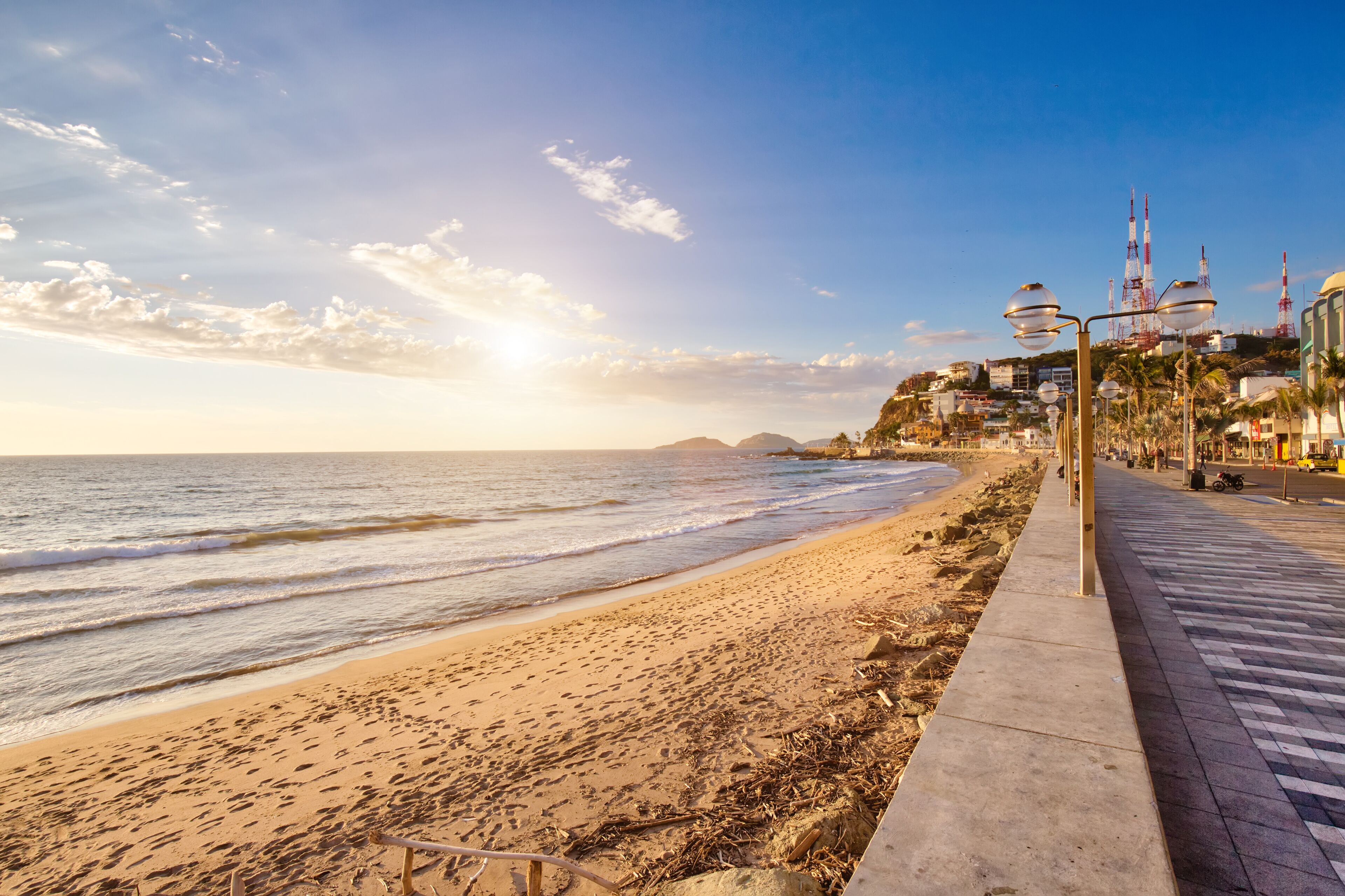 A beach with waves, a clear sky, and a cityscape with tall buildings and a rocket launch tower.