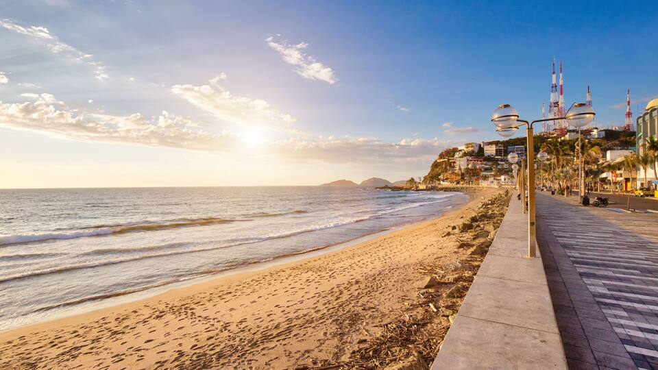 A beach with waves, a clear sky, and a cityscape with tall buildings and a rocket launch tower.