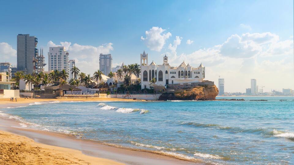 A coastal cityscape with a prominent white church, palm trees, and modern buildings.