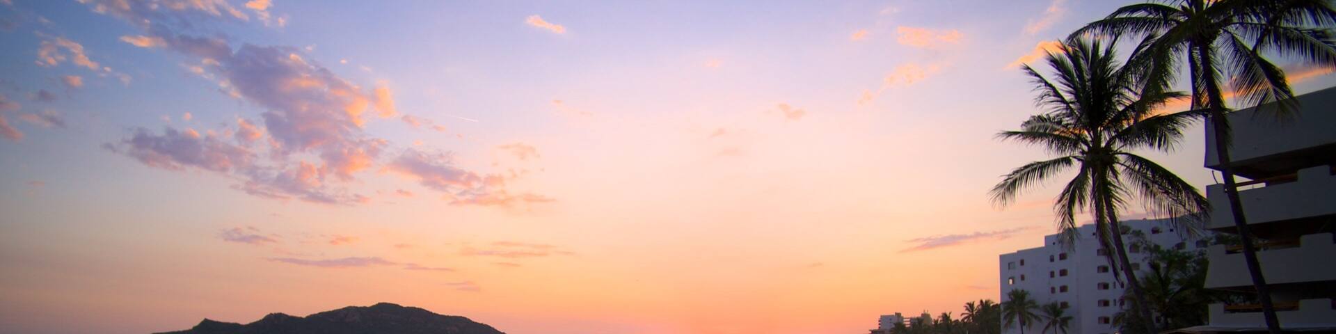 A beach at sunset with palm trees, ocean waves, and a distant mountain.