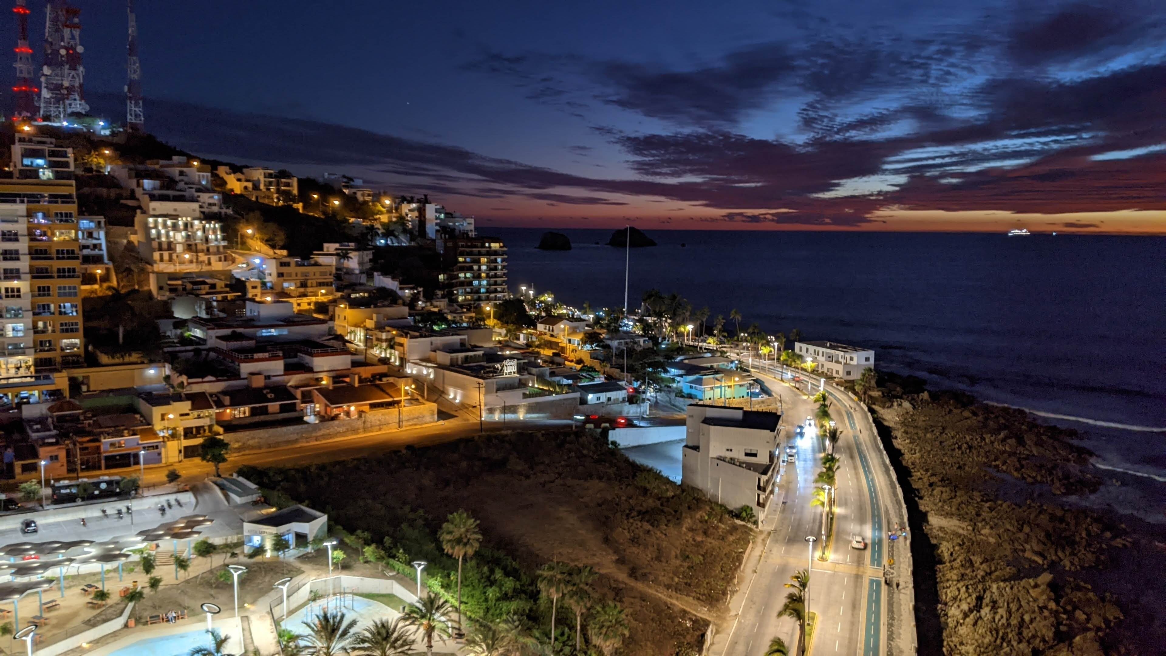 Un pueblo costero al atardecer con edificios iluminados, una carretera sinuosa y una playa.