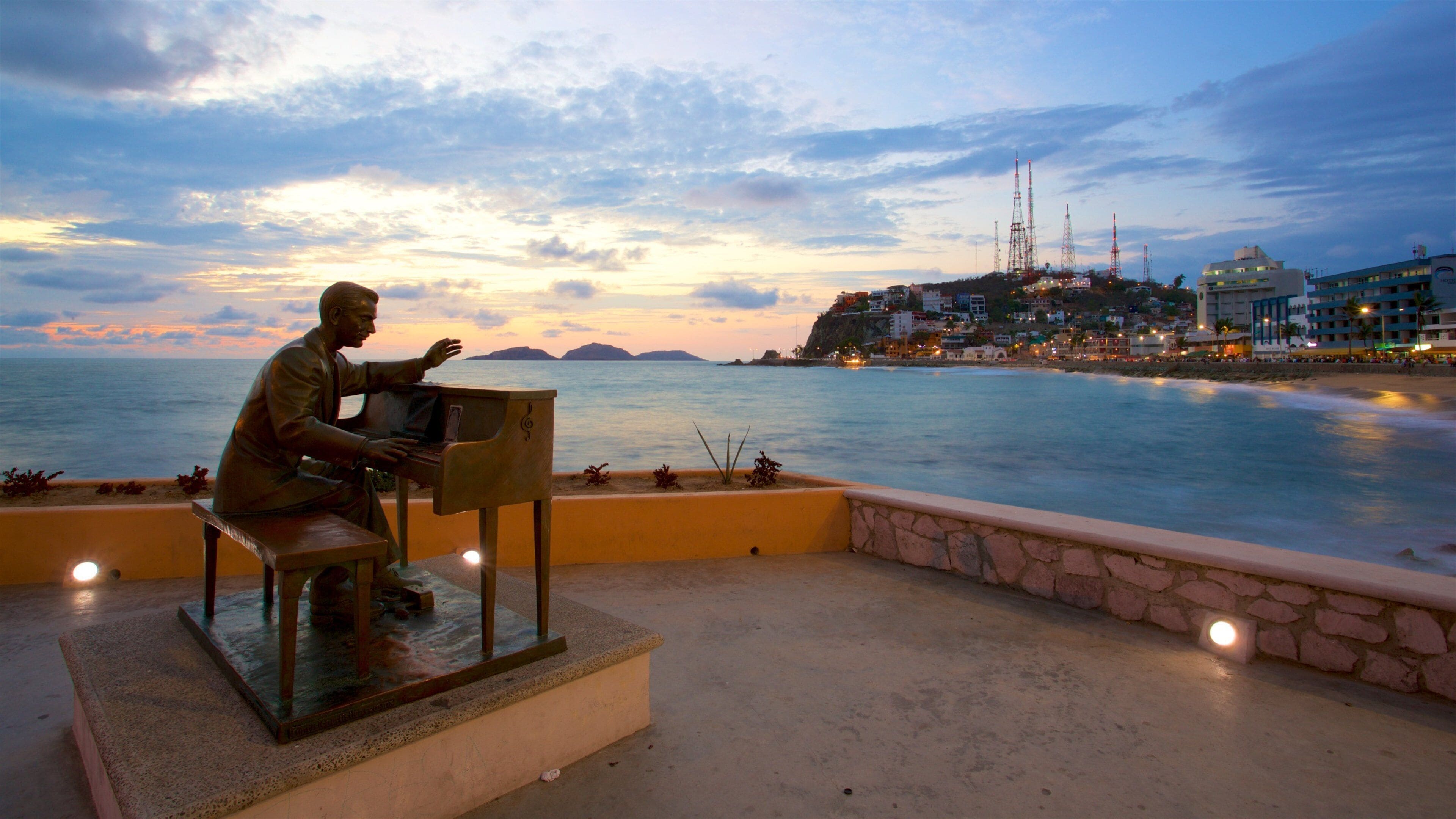 A statue of a man playing a piano overlooking a beach at sunset.