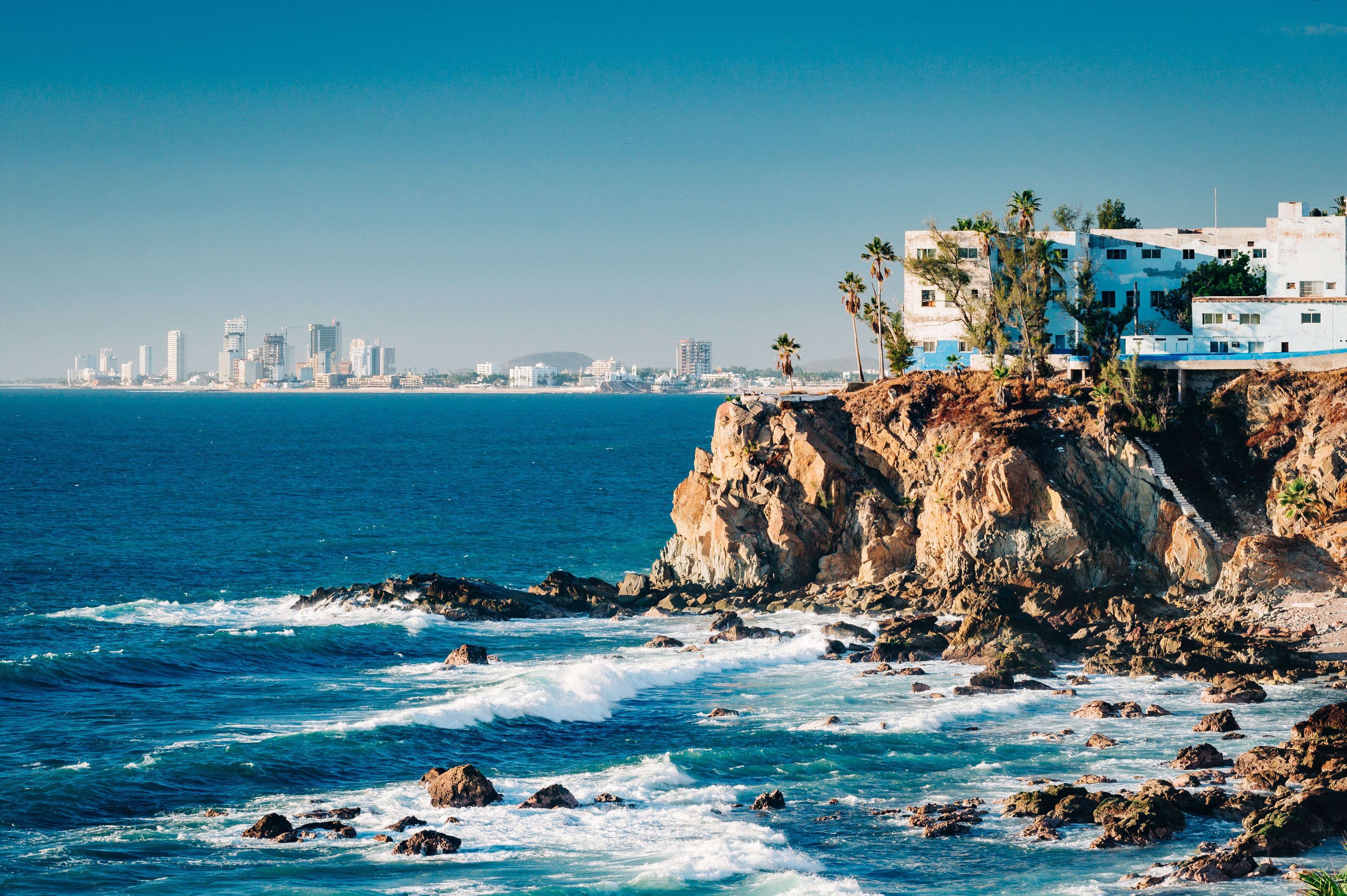 A coastal cityscape with a rocky shoreline, waves crashing against the rocks, and a city skyline in the distance.
