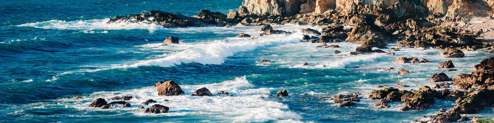 A coastal cityscape with a rocky shoreline, waves crashing against the rocks, and a city skyline in the distance.