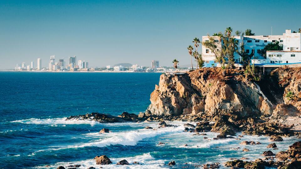 A coastal cityscape with a rocky shoreline, waves crashing against the rocks, and a city skyline in the distance.