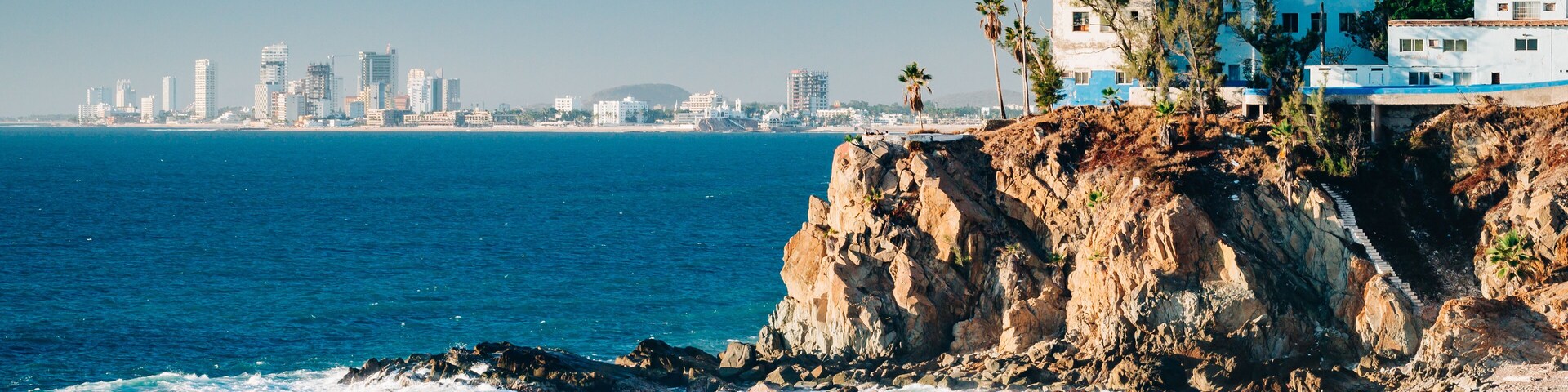 A coastal cityscape with a rocky shoreline, waves crashing against the rocks, and a city skyline in the distance.