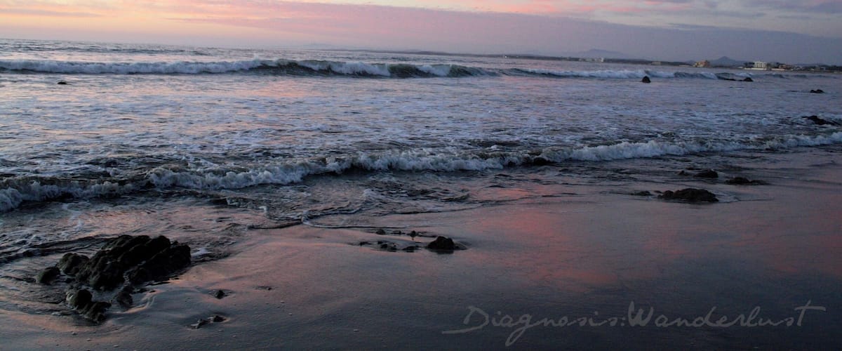 A beach at sunset with waves and a pink sky.