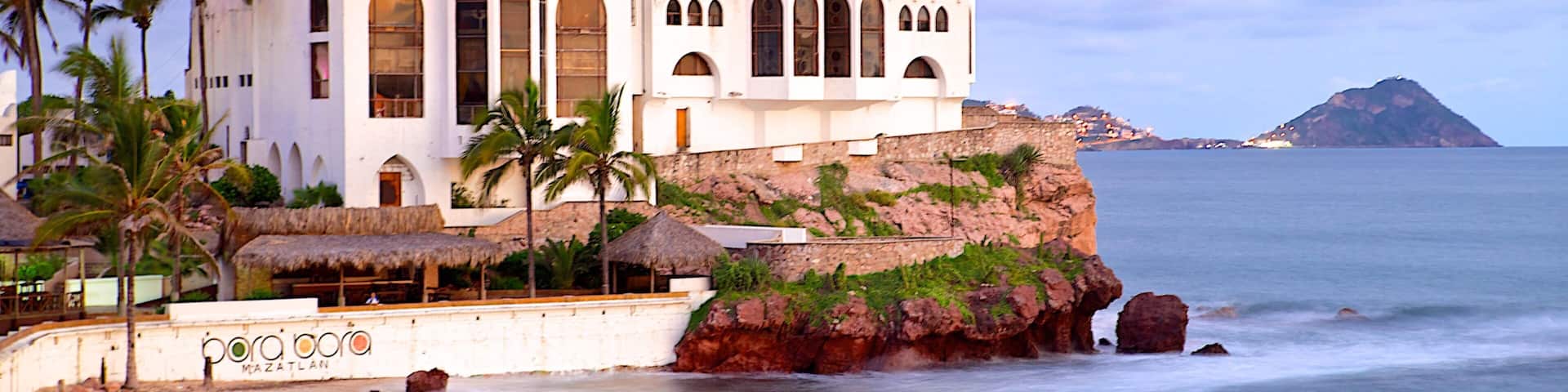 A white building with arched windows and green roofs near the ocean.