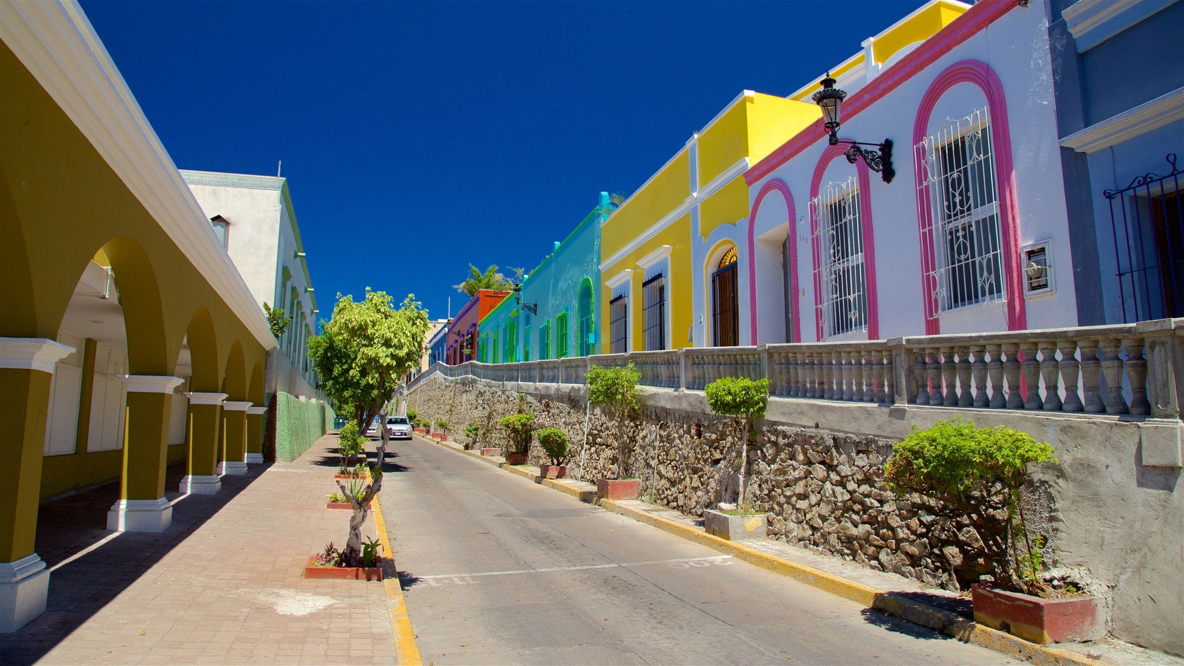 Una calle colorida bordeada de edificios, un muro de piedra y plantas en macetas.