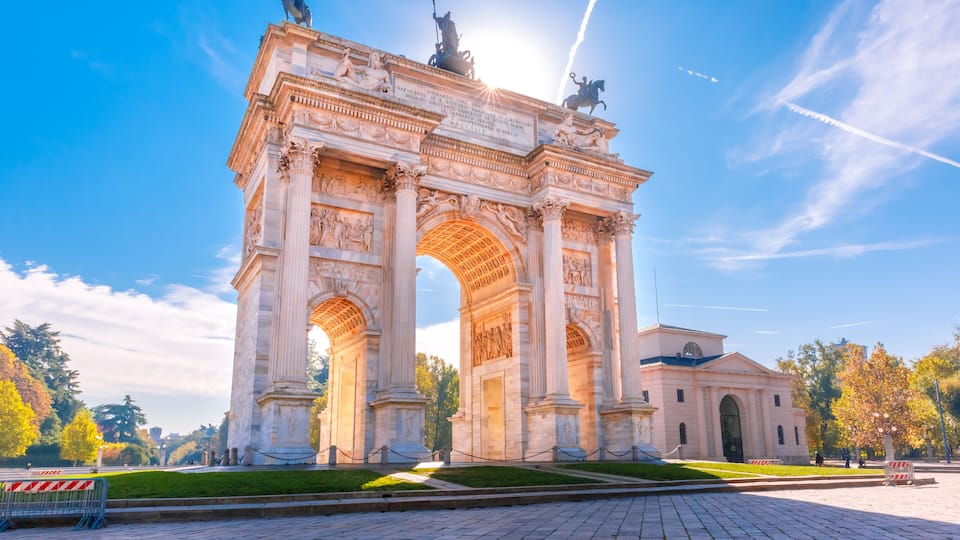 A large stone arch monument with statues on top, surrounded by a paved area and trees.