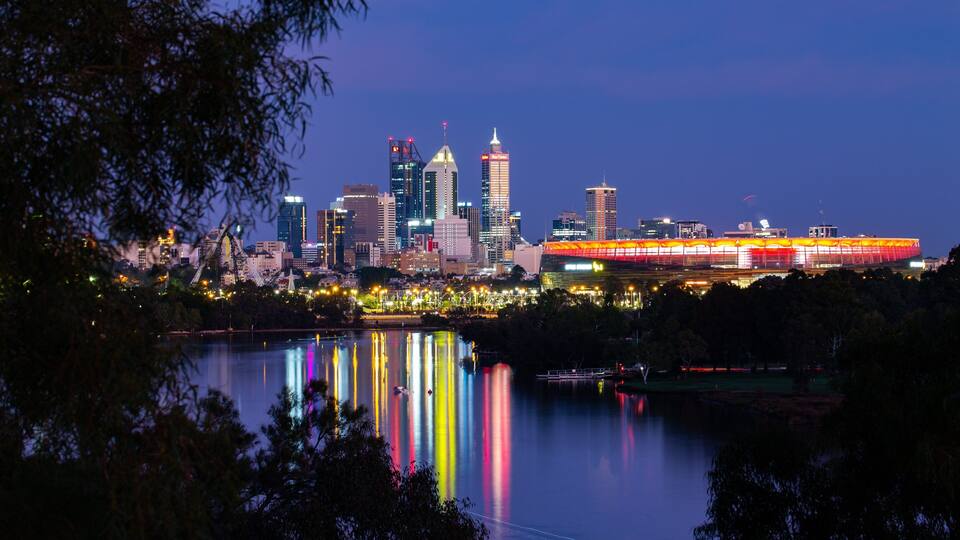 A city skyline at dusk with illuminated buildings and a stadium reflecting on the water.