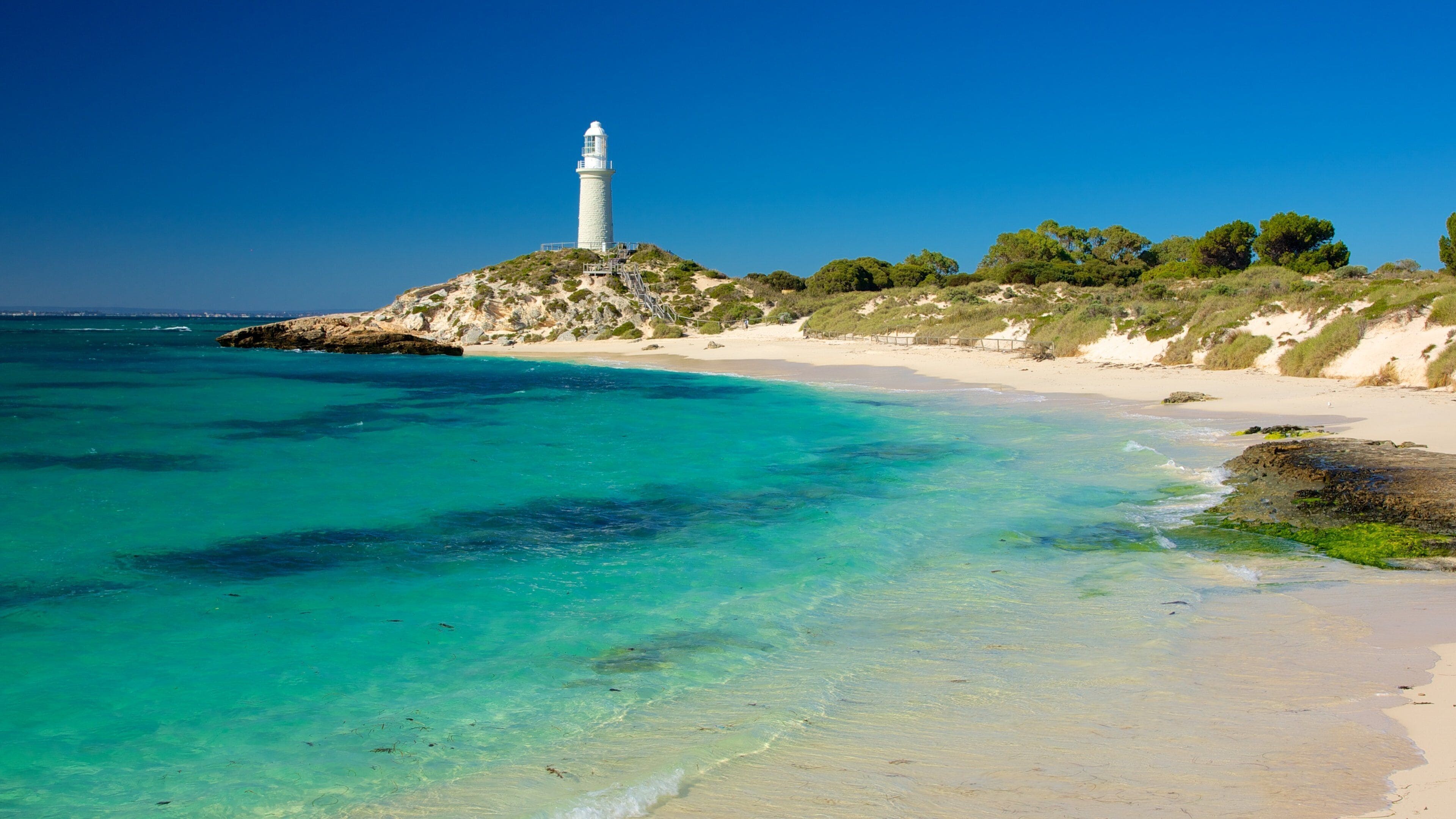 A lighthouse on a rocky outcrop, clear turquoise waters, and a sandy beach.