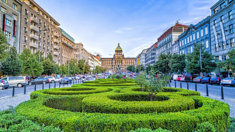 A city square with a large building in the background, surrounded by neatly trimmed hedges and parked cars.