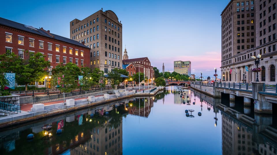 A calm river reflecting buildings, a bridge, and a historic building with a clock tower.