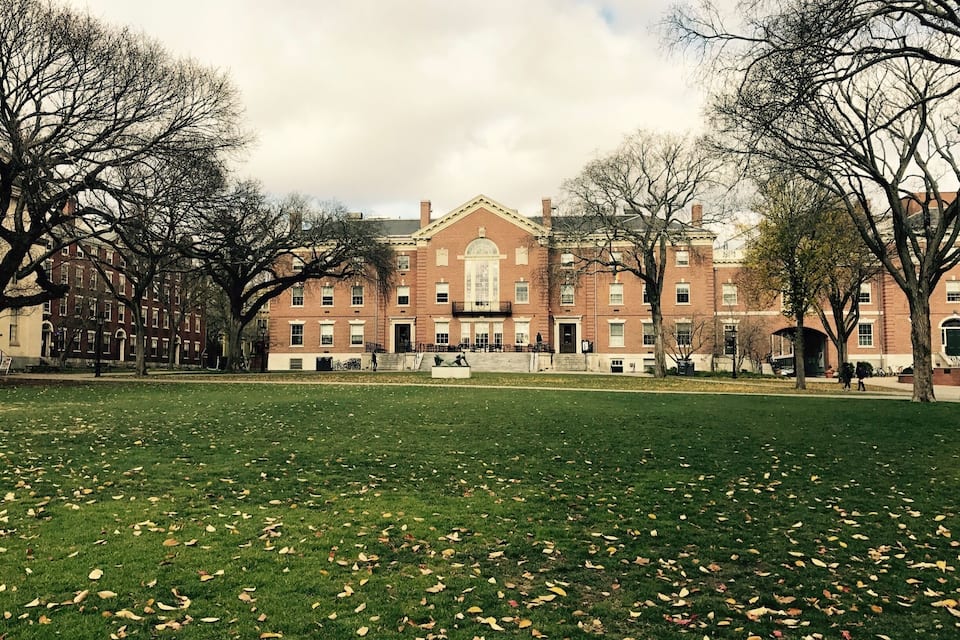 A large brick building with a central entrance, surrounded by a grassy field and leaf-covered ground.