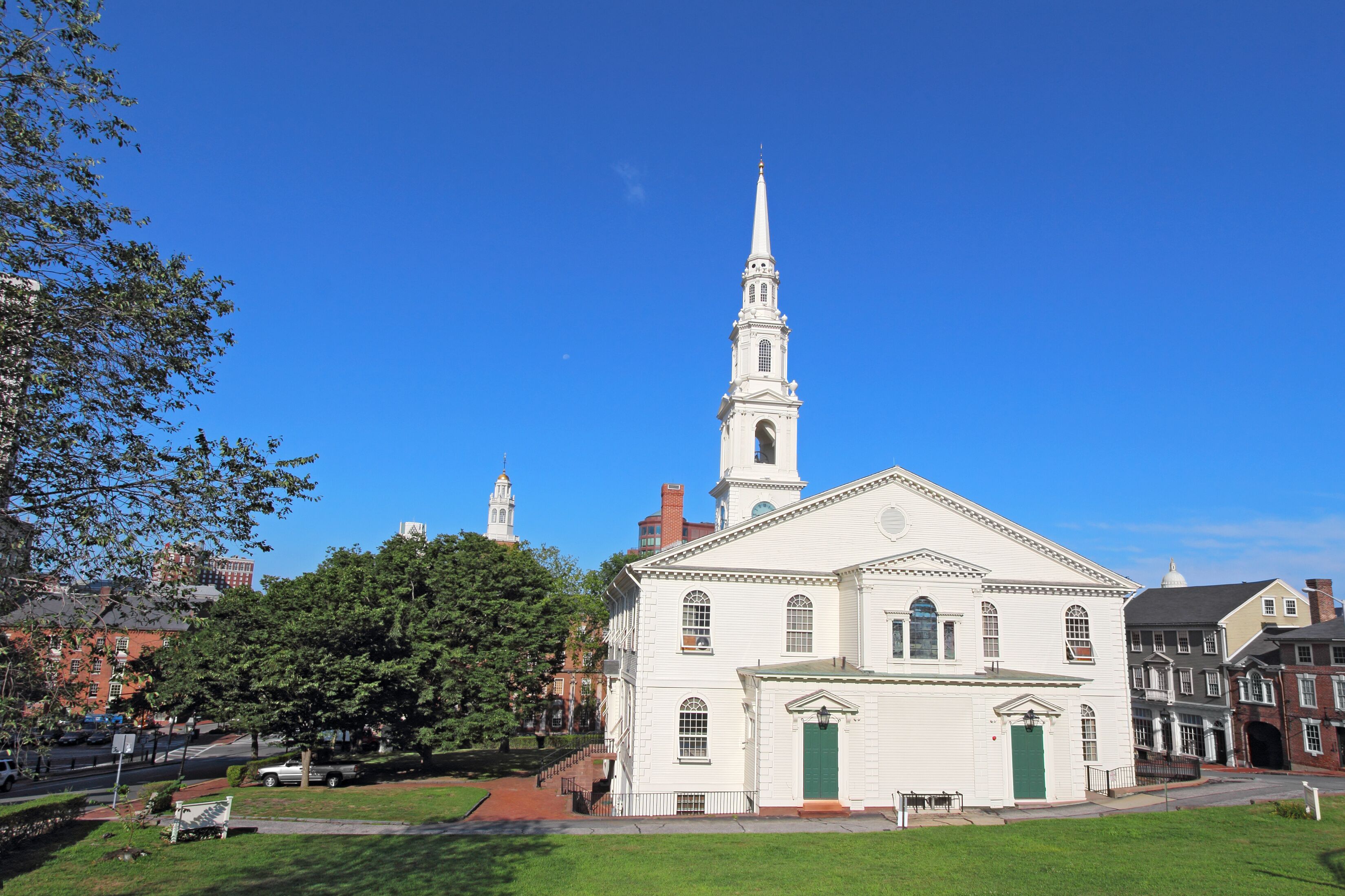 A white church with a tall steeple and a green door, surrounded by trees and other buildings.
