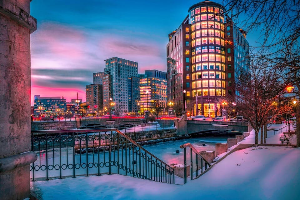 Escena urbana nevada con un río, un puente y edificios iluminados al anochecer.