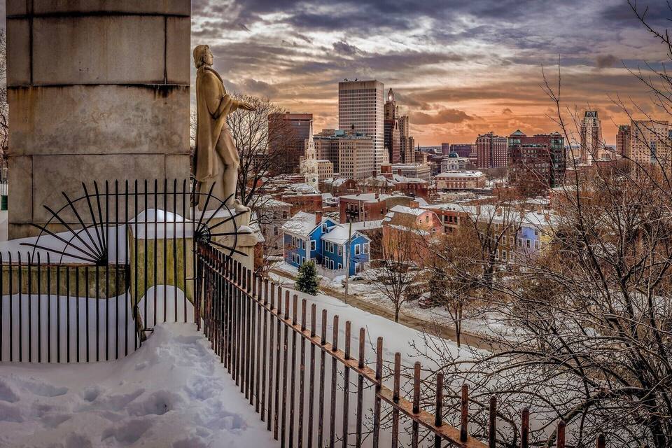 A cityscape with a statue, snow-covered buildings, and a cloudy sky.