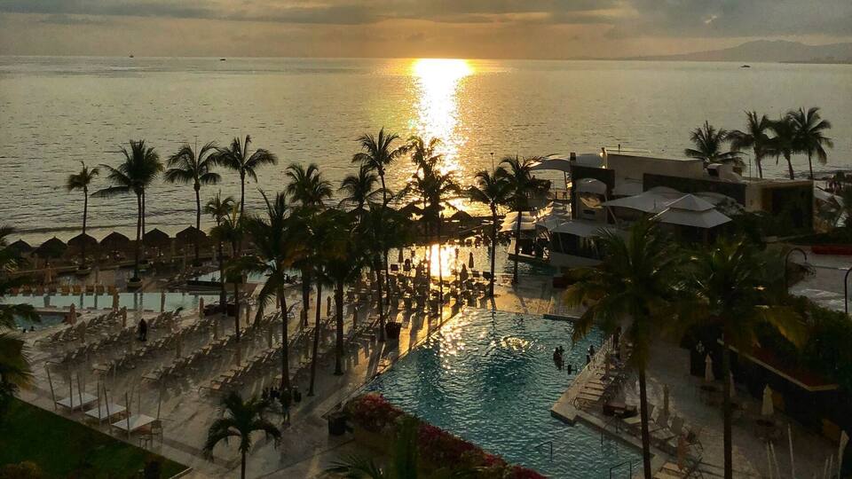 A sunset over a beach with a pool and palm trees.