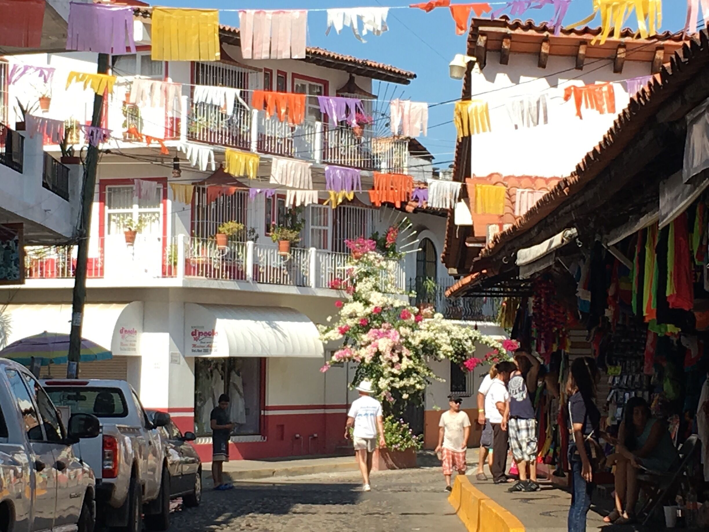 A street with colorful laundry hanging from balconies, parked cars, and a flower display.